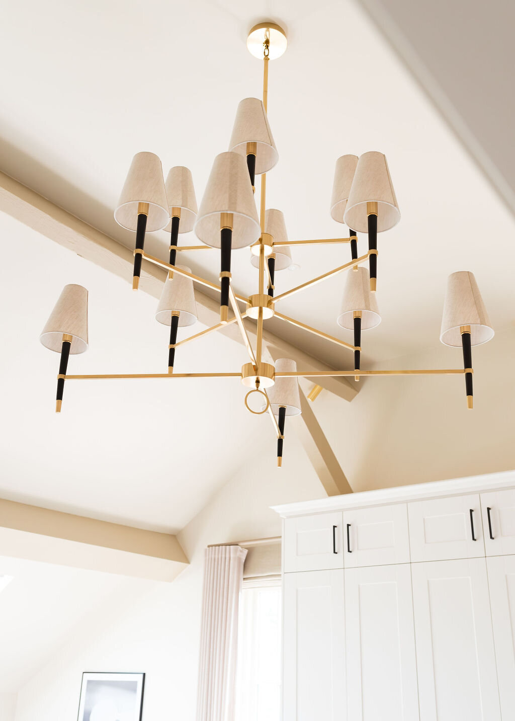 Modern chandelier with fabric-covered light shades hanging from a high, vaulted ceiling. The background includes white cabinetry and a partial view of a framed picture on the wall. The overall decor is minimalist and elegant.