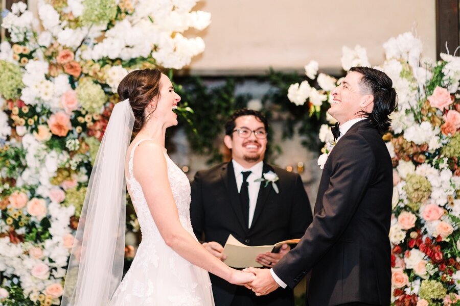 A heartfelt exchange of laughter and vows between a bride and groom framed by a breathtaking floral installation. Their radiant smiles and genuine joy light up the altar in this unforgettable moment.