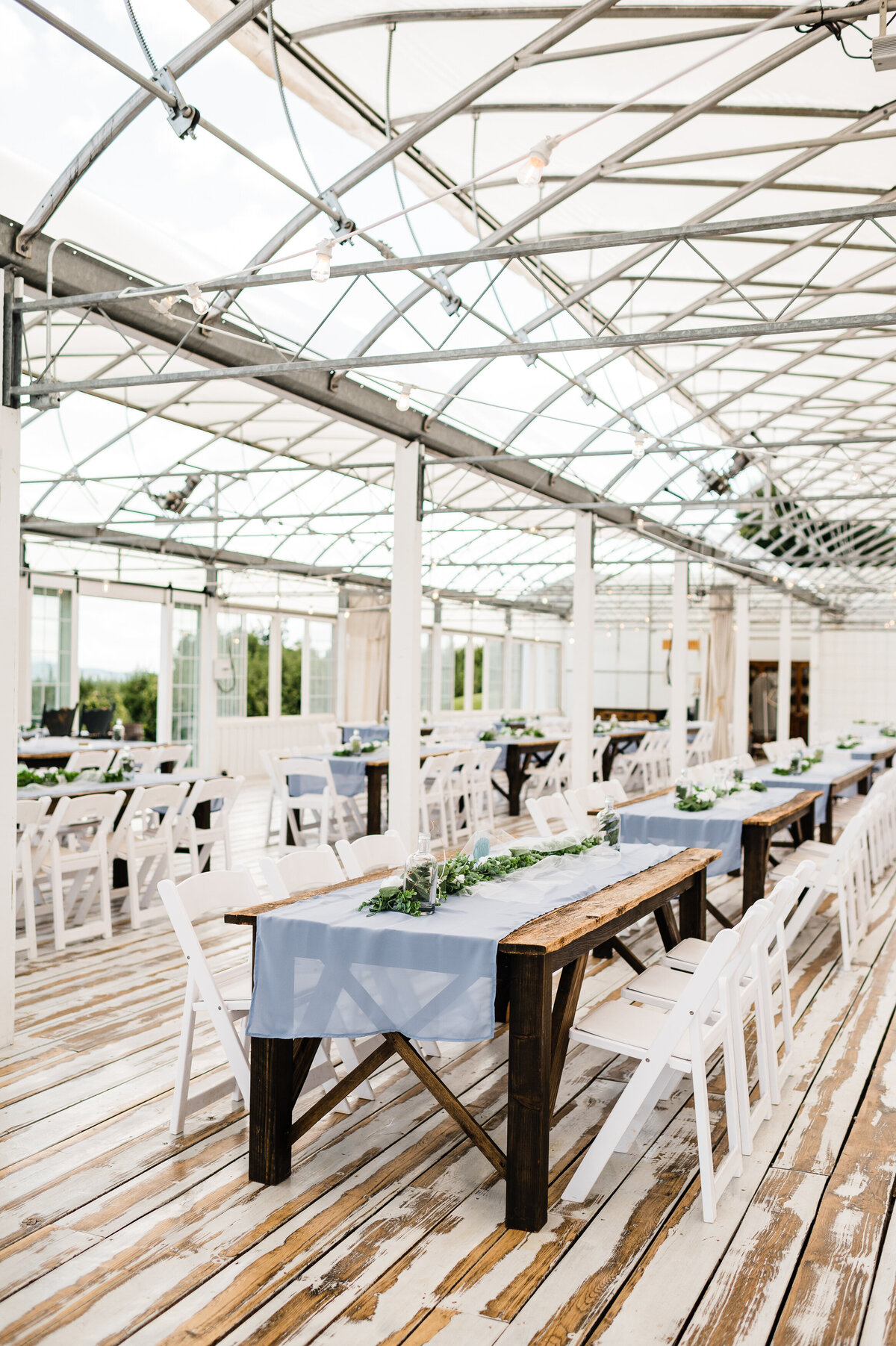 greenhouse wedding venue decor with dark wood tables and white chairs decorated with light blue table runners and greenery down the center