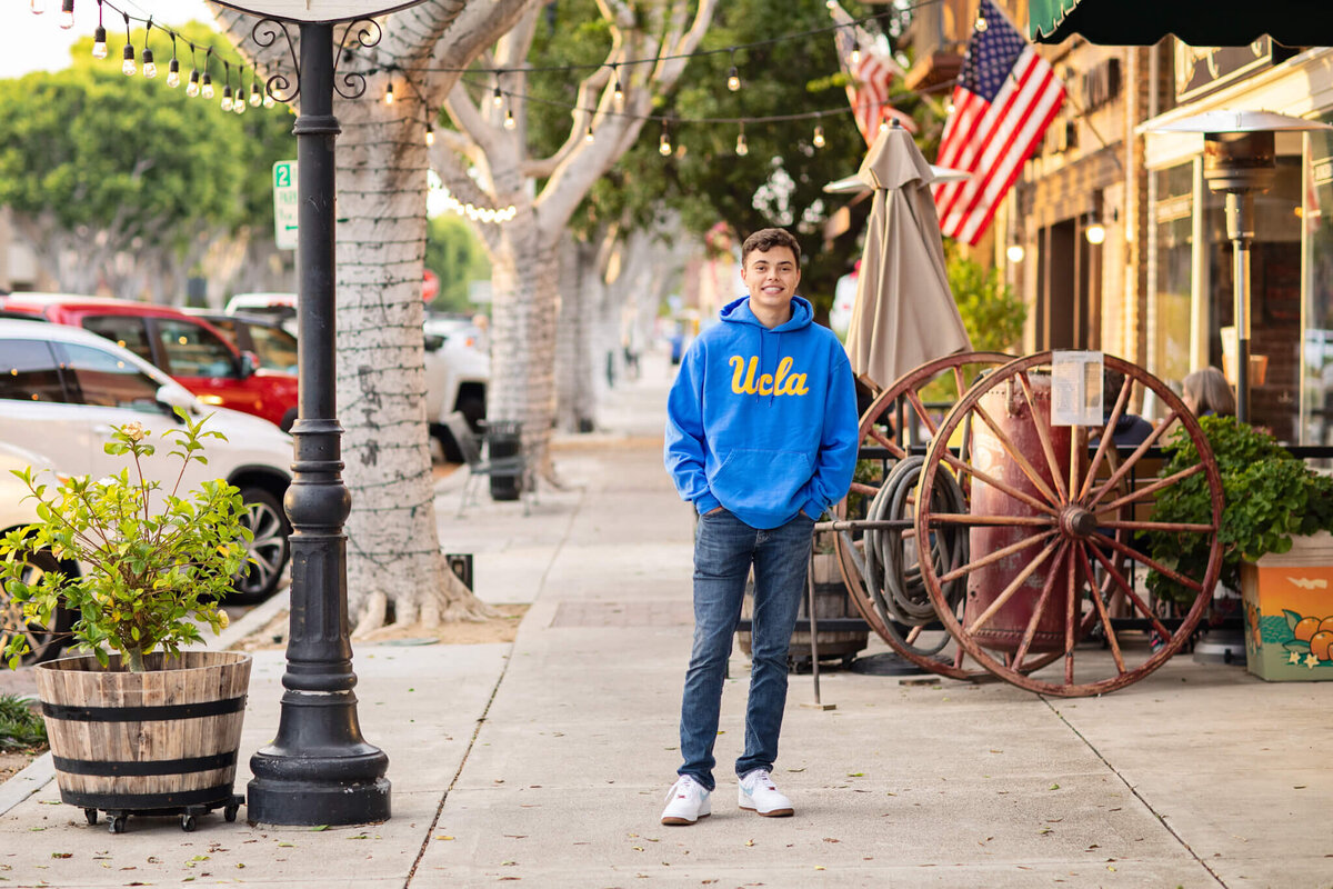 A person in a blue UCLA hoodie stands on a sidewalk with hands in pockets. They are surrounded by trees, parked cars, and shops, with an American flag hanging nearby. A large wagon wheel is visible against a brick wall.