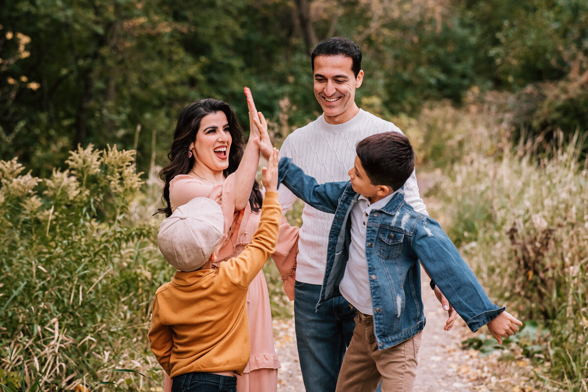 A family of four in a moment of celebration, with the mother and sons high-fiving each other while the father looks on, all smiling brightly amidst a natural, wooded setting.