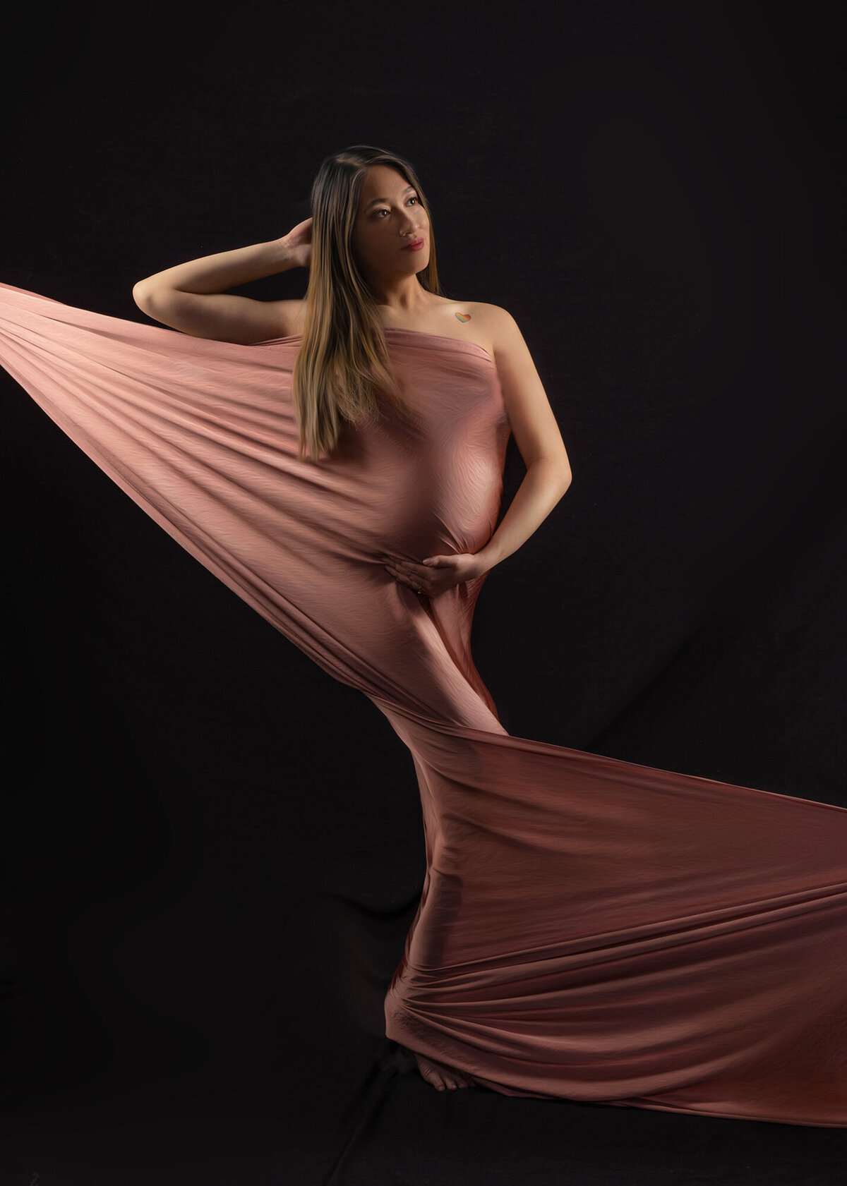 An image from the studio of an Iowa maternity photographer of a pregnant woman wrapped in pink sheets being pulled in both directions with a hand on her bump