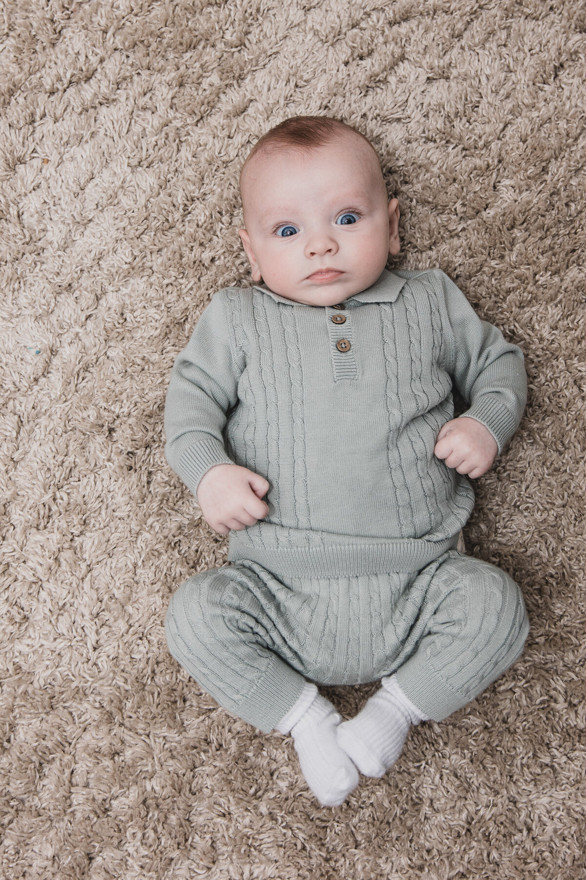 Baby in a gray knitted outfit lying on a fluffy beige carpet, looking upwards with wide blue eyes.