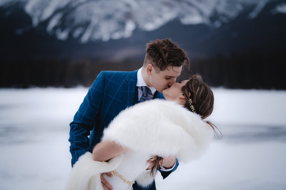 Wedding couple dip and kiss in front of frozen lake in Jasper, Alberta