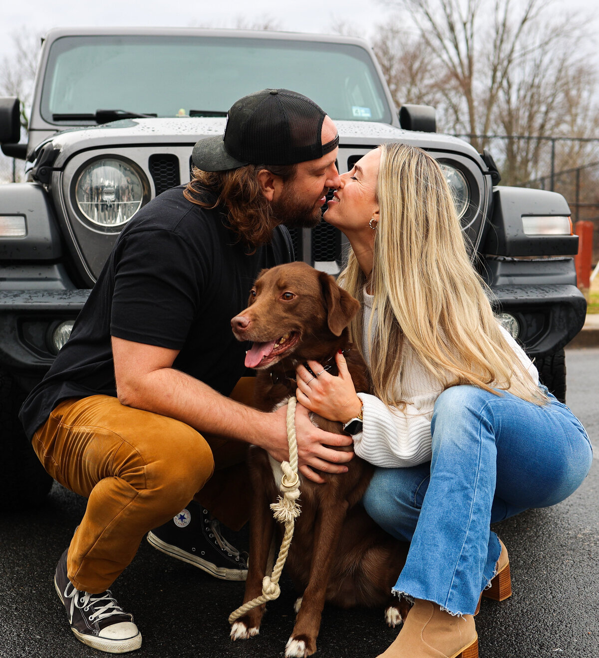 man wearing hat backwards and woman wearing jeans kissing with brown dog between them