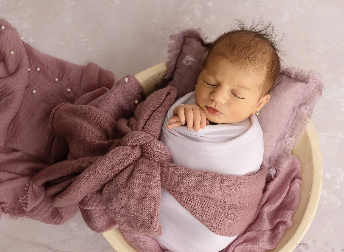 Baby girl in white and mauve swaddles is caputred for her brooklyn ny newborn photoshoot Her hands are peeking out of the swaddle. Captured by top Brooklyn newborn photographer Chaya Bornstein