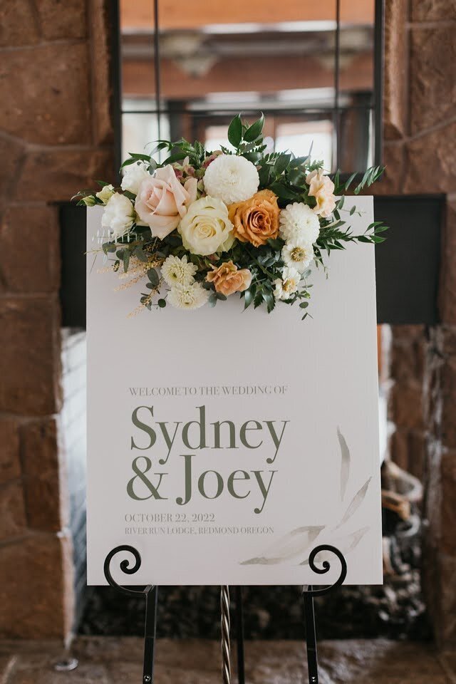 Wedding welcome sign with floral arrangement sitting atop it.