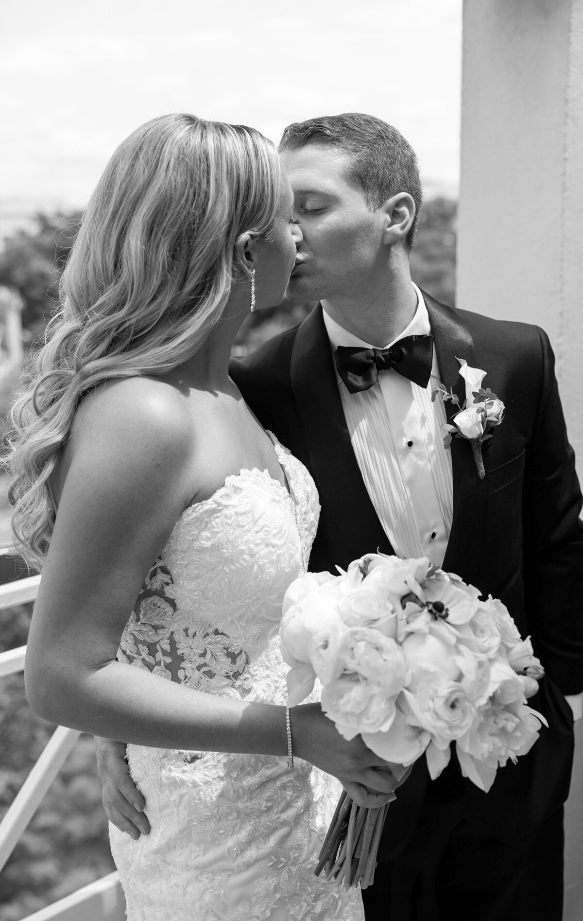 A bride and groom kiss on a balcony. The bride wears a lace wedding dress and holds a bouquet of white flowers. The groom wears a tuxedo with a bow tie and boutonniere. The setting is bright and outdoors.