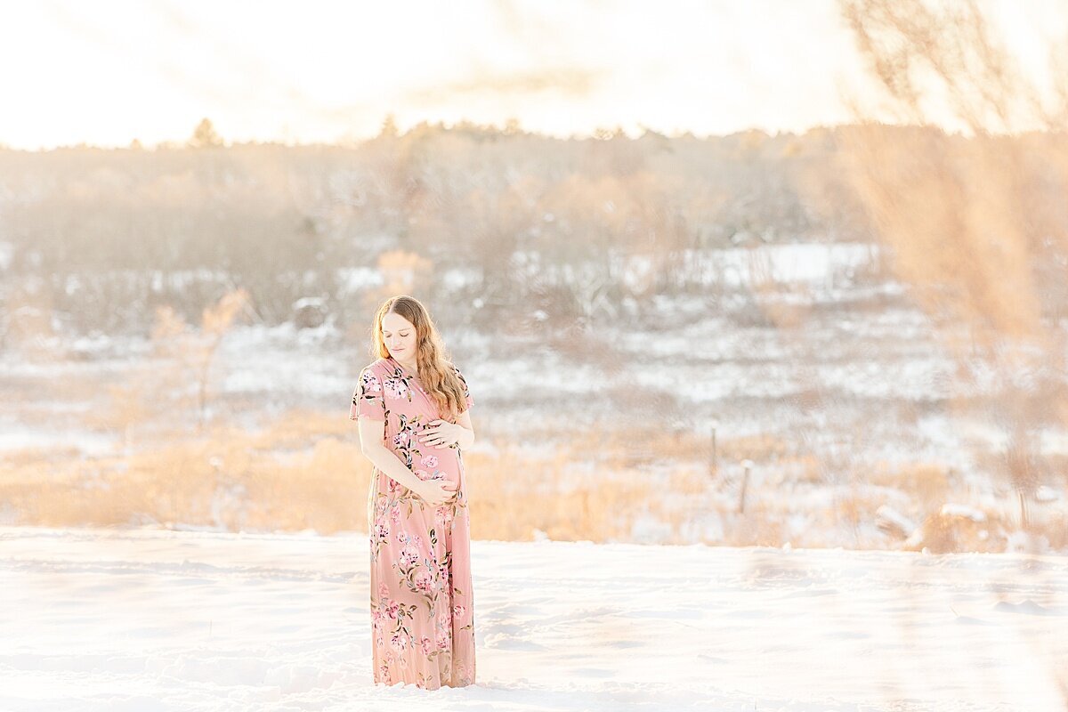 pregnants womand stands in field during snowy winter maternity photo session with Sara Sniderman Photography in Medfield Massachusetts