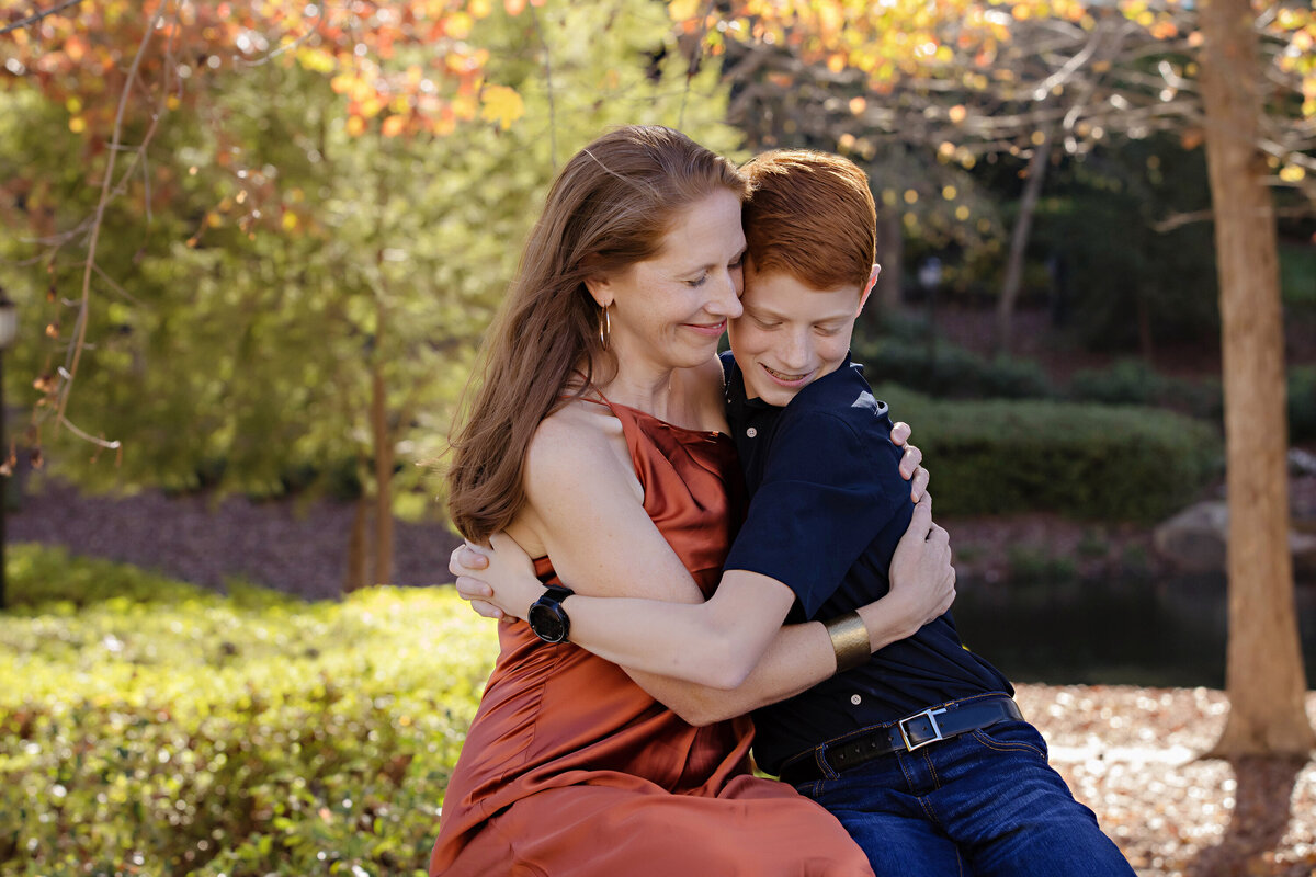 Cozy autumn portrait of mom and son seated together on park bench surrounded by fall foliage at Ballantyne Corporate Park