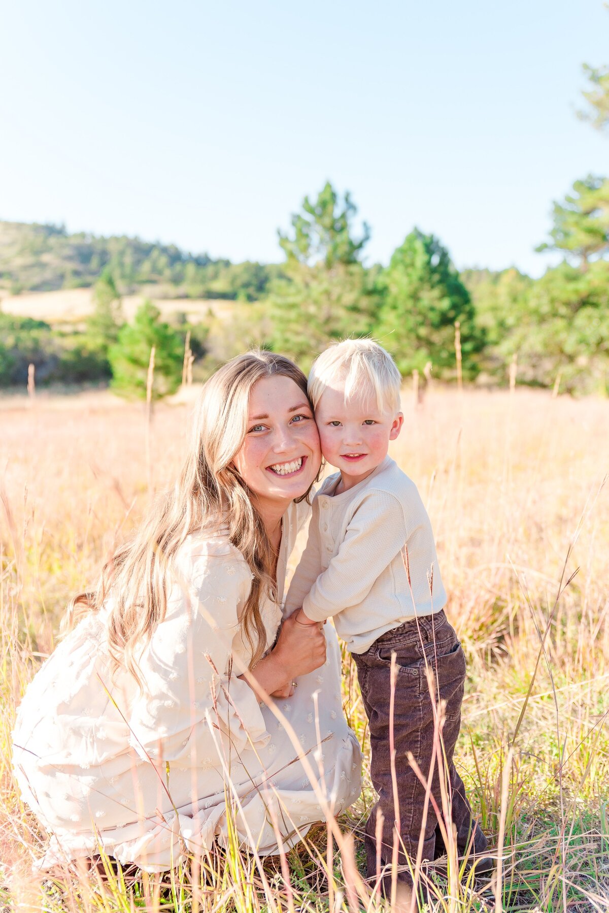 A mom in a long sleeve tan dress holds her son's hands and leans in toward him as he stands and they both smile towards the camera held by denver family photographer