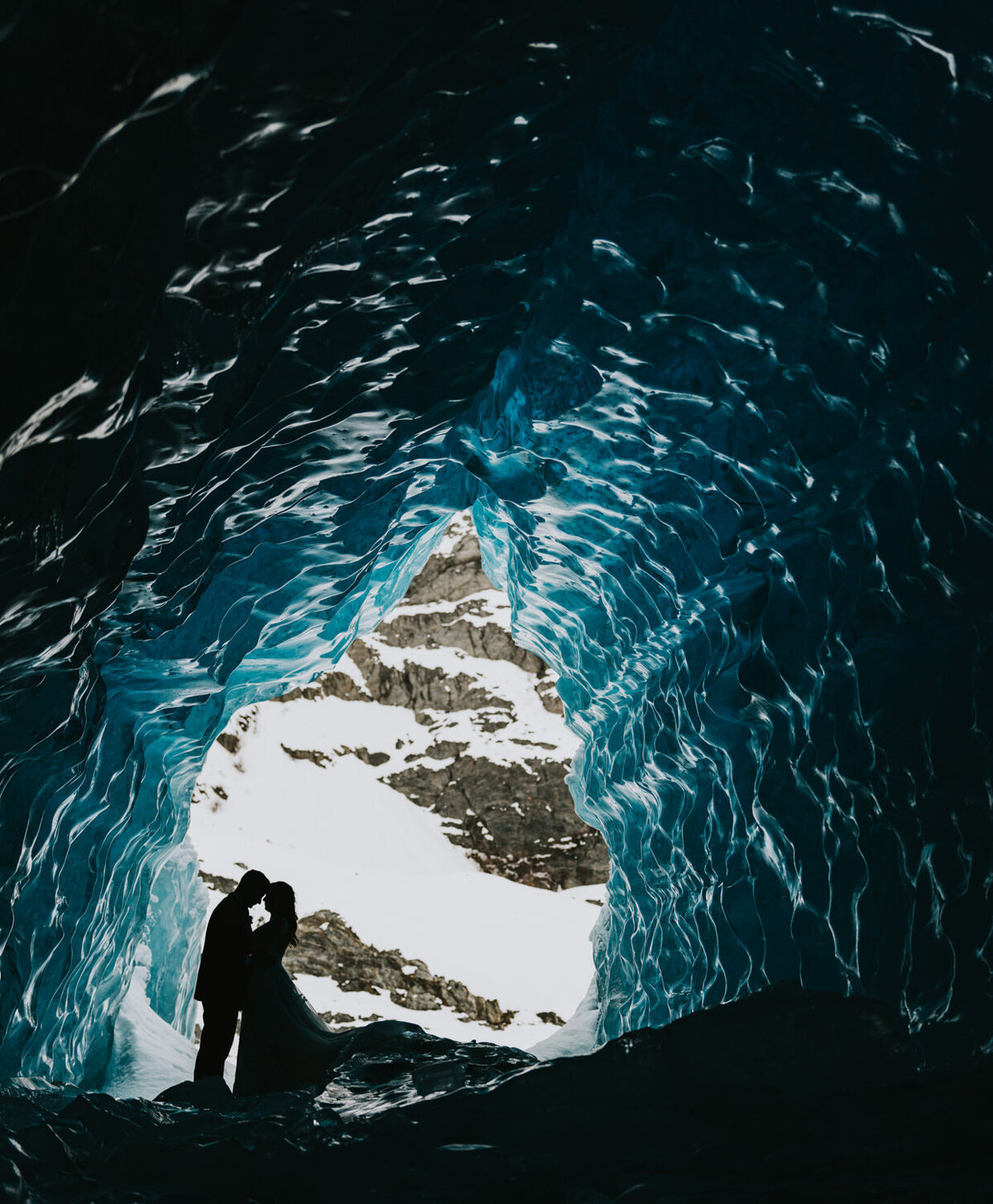 a couple standing in a striking blue ice cave on a glacier in alaska in their wedding attire on their elopement day