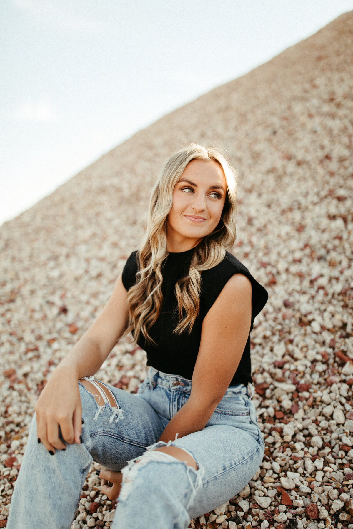 Girl senior wearing black shirt and blue jeans sitting down and posing at gravel pit
