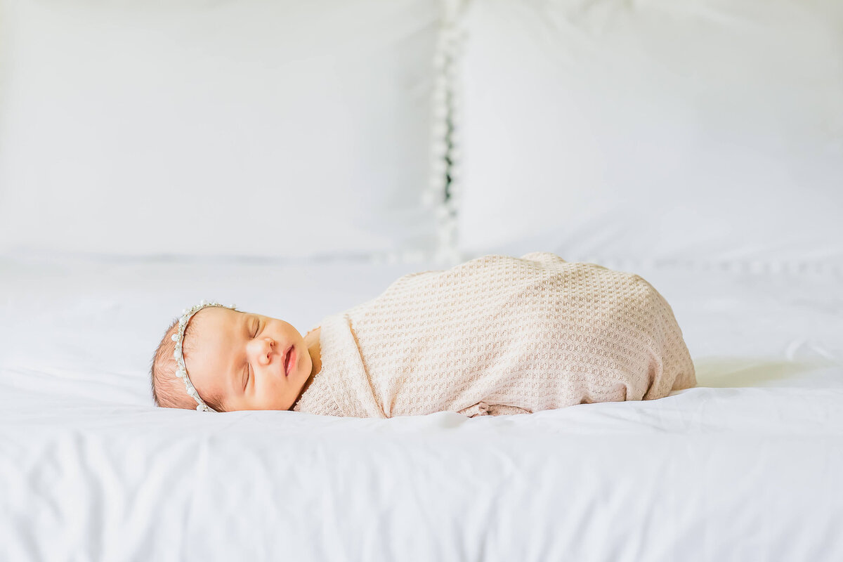 Newborn baby girl in a cream wrap and pearl bow laying on a bed with a white boho comforter