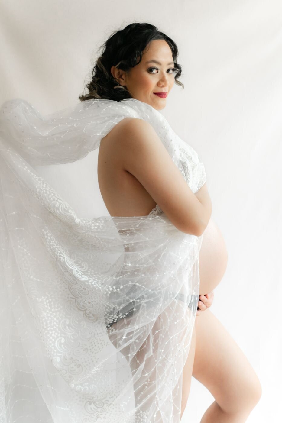 Expecting mom wrapped in a sheer white lace fabric, smiling and gently cradling her belly.