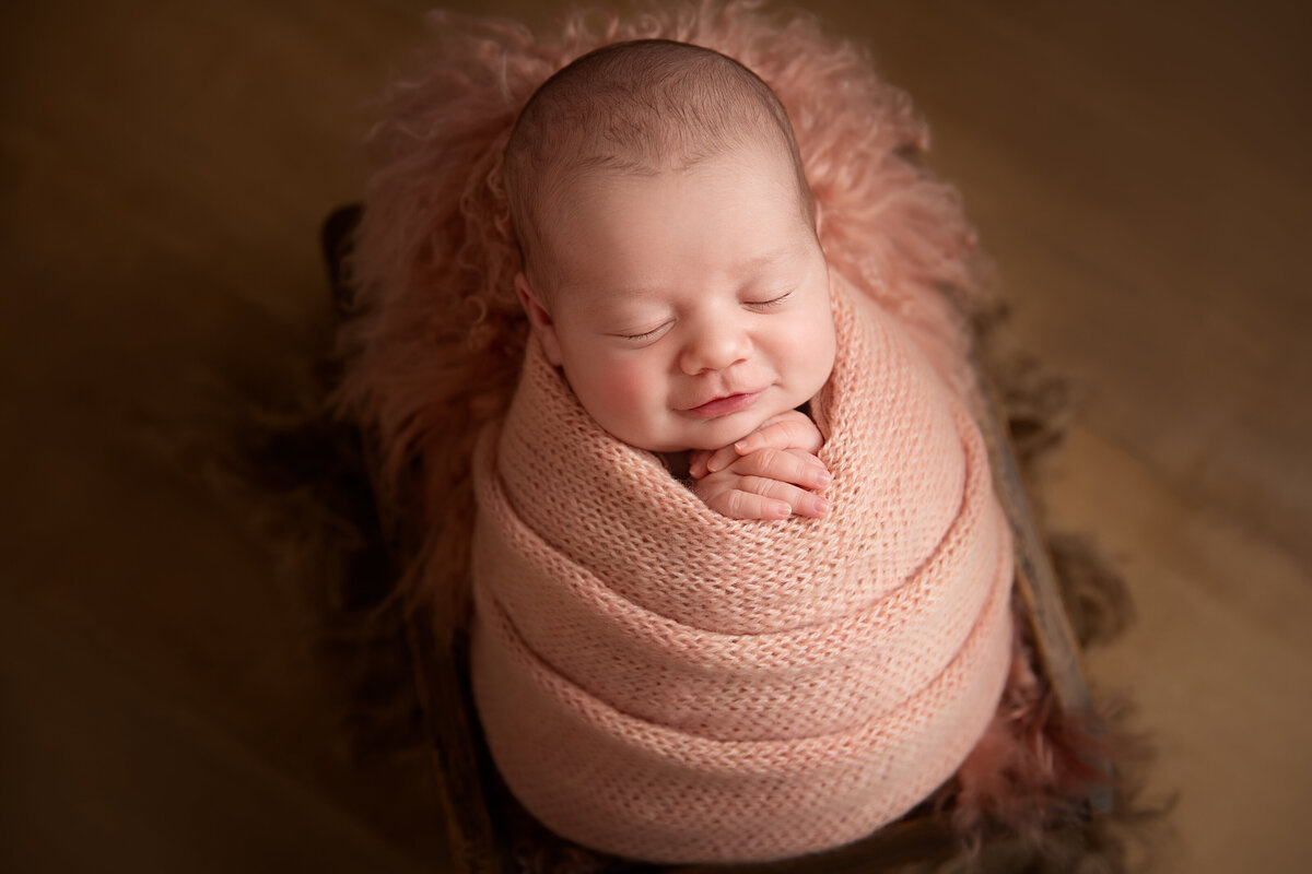 Newborn baby wrapped in a pink blanket on matching fur in a wooden box with a smile on her face.