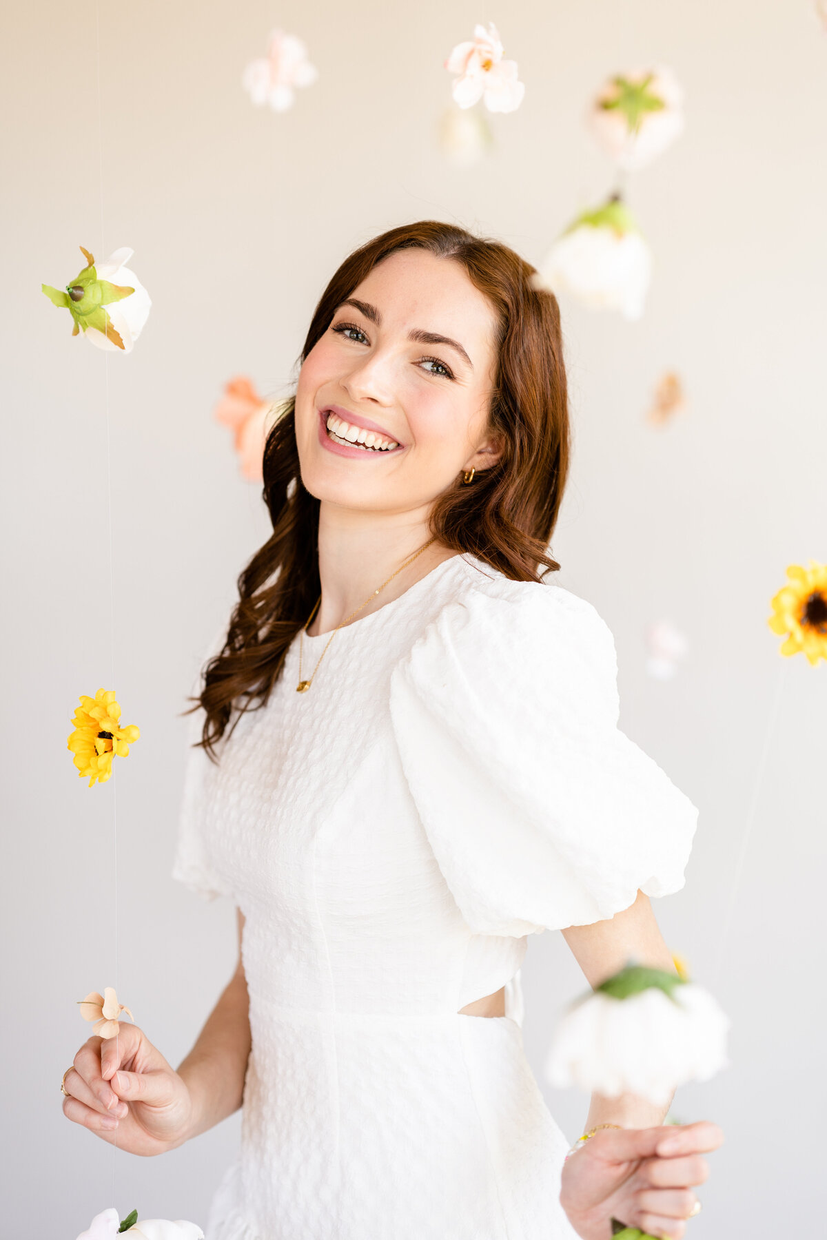 Senior girl wearing white dress and laughing at camera in the middle of hanging flowers at Bravely Studio
