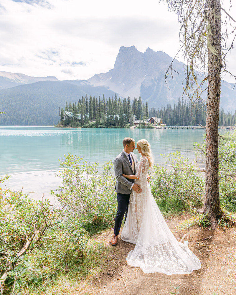 This romantic photograph captures the groom holding the bride as they face each other on the serene shoreline of Emerald Lake Lodge in Yoho National Park. The calm waters and majestic surroundings add a timeless, intimate feel to their special moment.