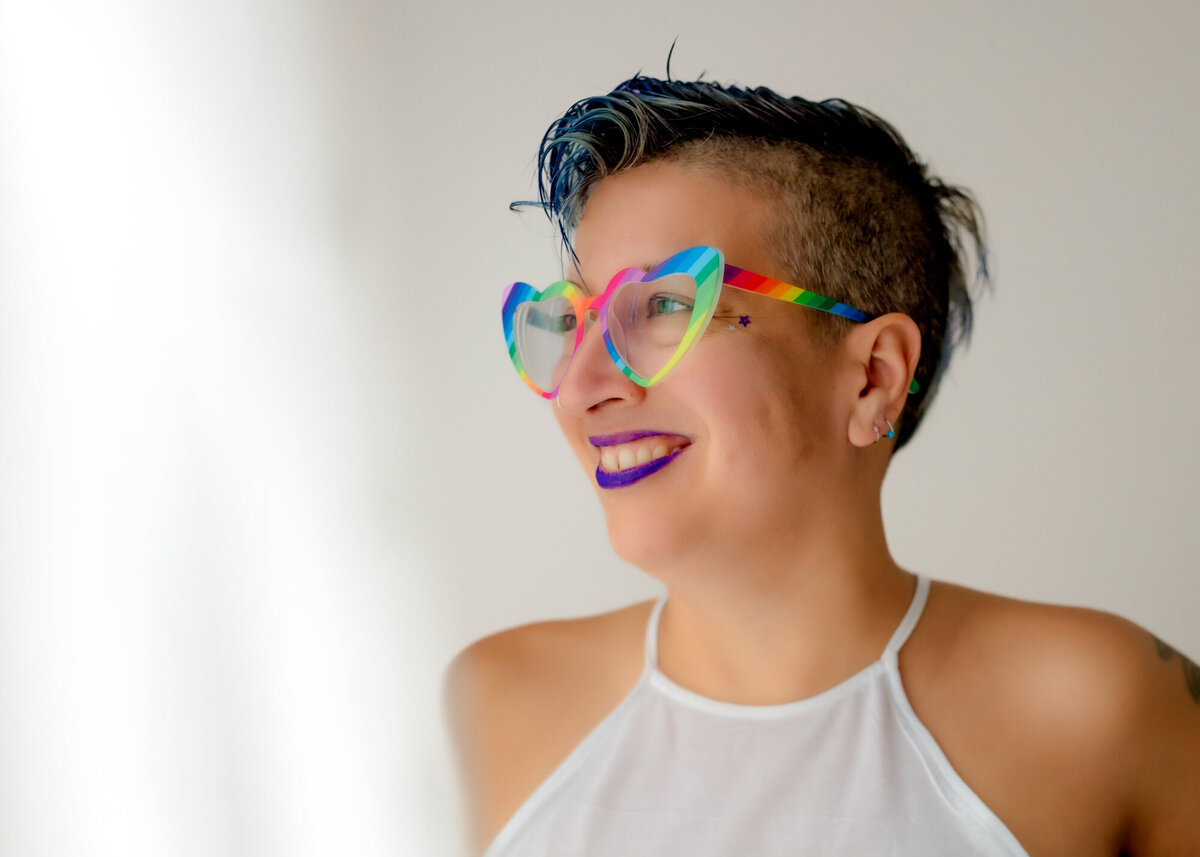 They/them headshot client wearing a white top, rainbow glasses and smiling out the window