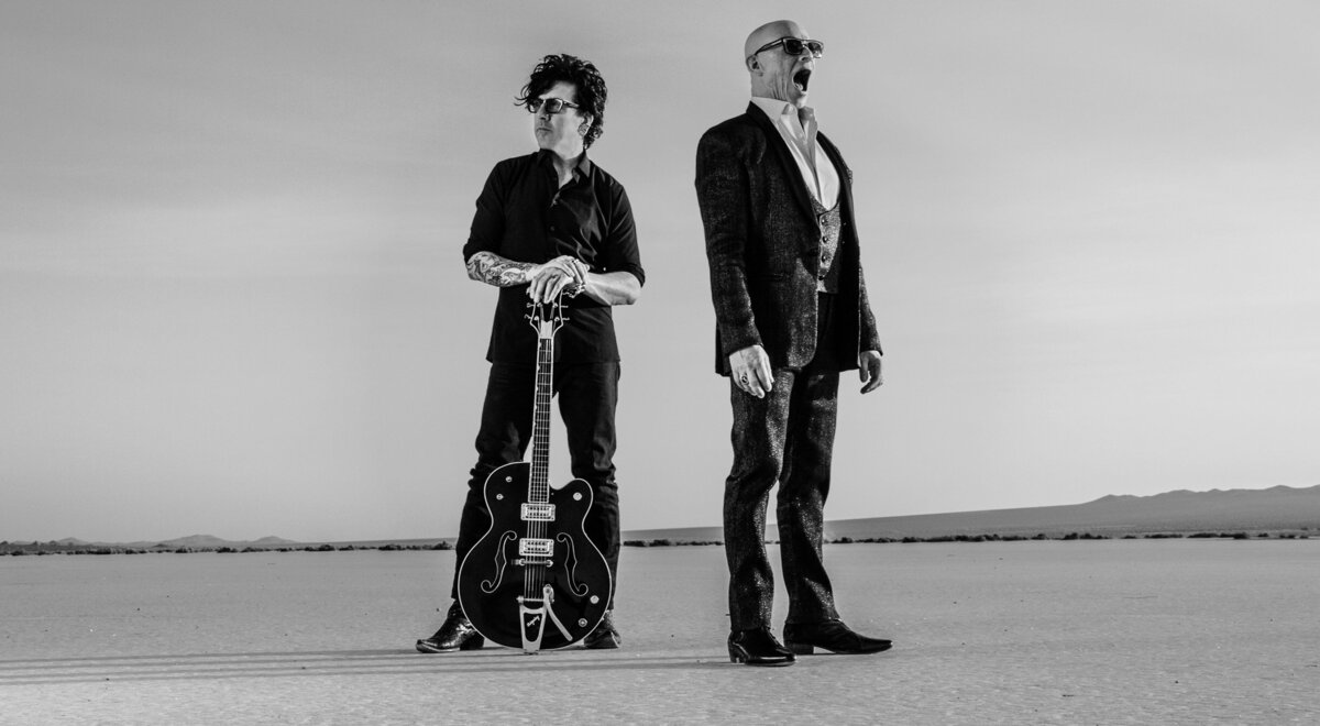 Black and white band portrait horse duo standing in desert one member has hands resting on neck of huitar standing upright between his leg singer drummer looking away with mouth open El Mirage