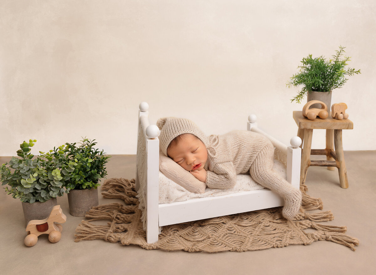 Brooklyn newborn photoshoot. Baby is sleeping on his belly on a miniature prop bed with one leg draping over the side of the bed. Baby's hands are resting under his chin. faux plants surround the baby. Captured by top Brooklyn newborn photographer Chaya Bornstein