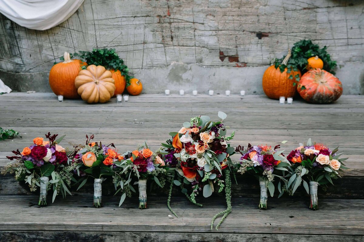Several vibrant floral bouquets featuring roses, eucalyptus, and other colorful flowers are lined up on wood stairs. Pumpkins and candles decorate the background.