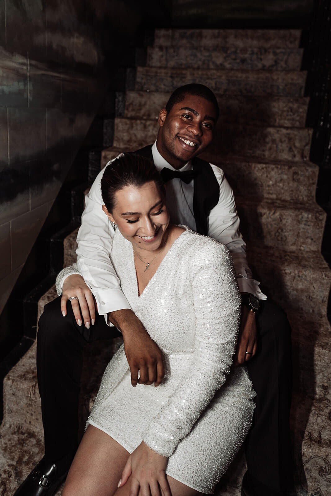 groom drapes his arm over the bride as they sit laughing on the stairs at the nomad hotel london at their intimate wedding party