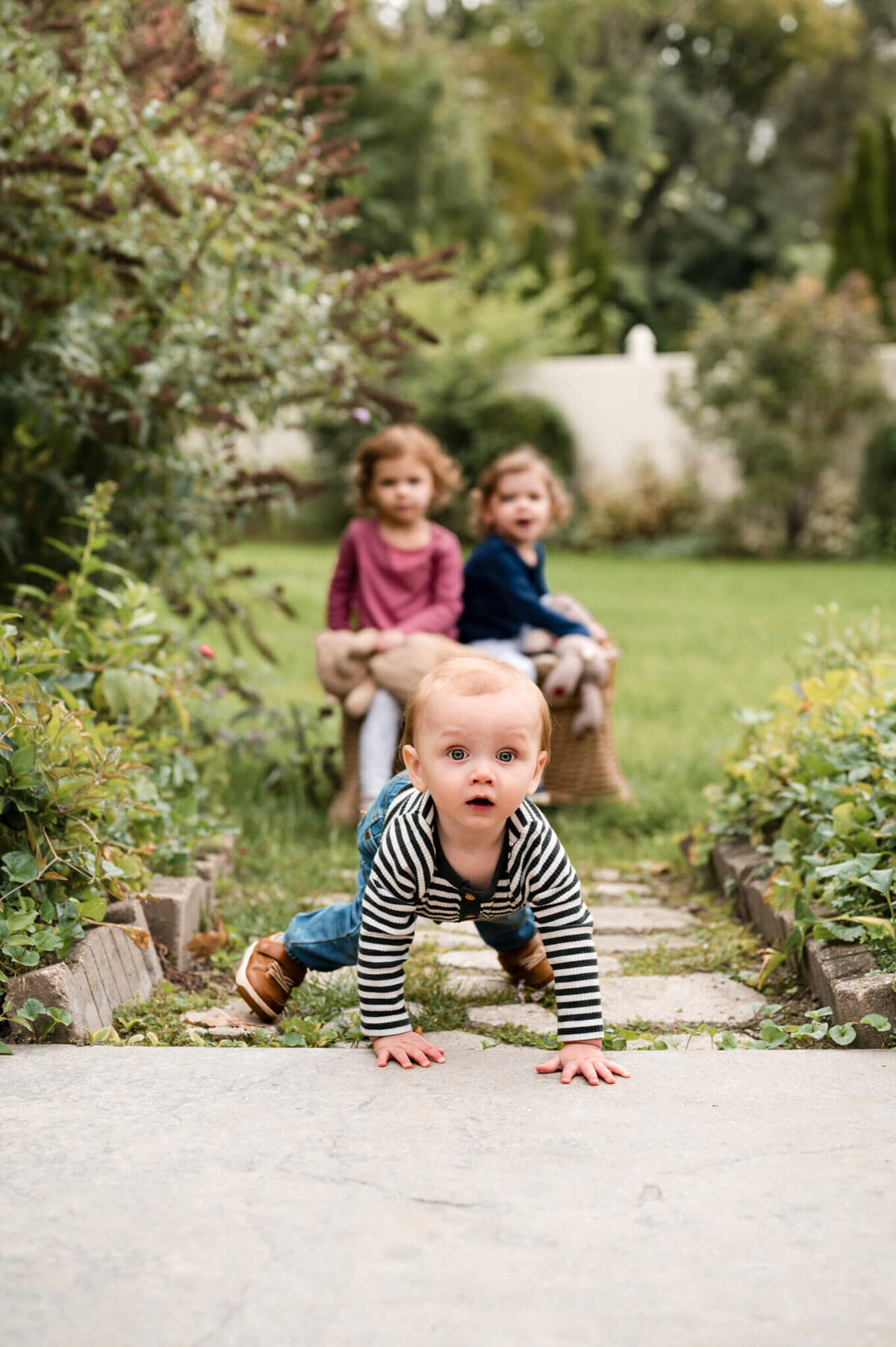 Young baby is looking and crawling toward the camera, while older siblings are blurred in the background looking at him.