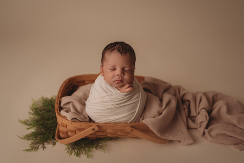 Newborn session with Atlanta, Georgia newborn photographer. 2 week old baby boy at atlanta's best newborn photography studio posed in a cream swaddle wrap sitting in a woven basket with creams and greens.