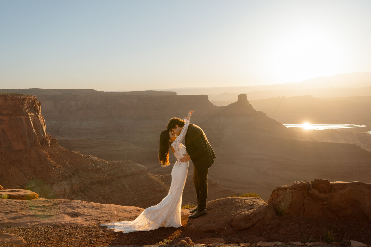 Bride and groom on the cliffs of Moab during sunrise embracing each other with her Claire Pettibone Shenandoah long sleeve lace gown