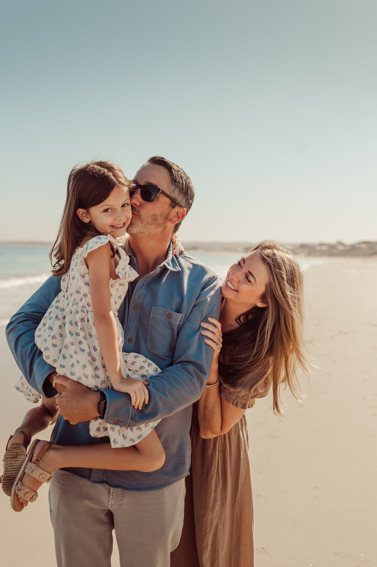 Dad kissing his young daughter's cheek while holding her on a sunny Monterey beach, mom smiling beside them