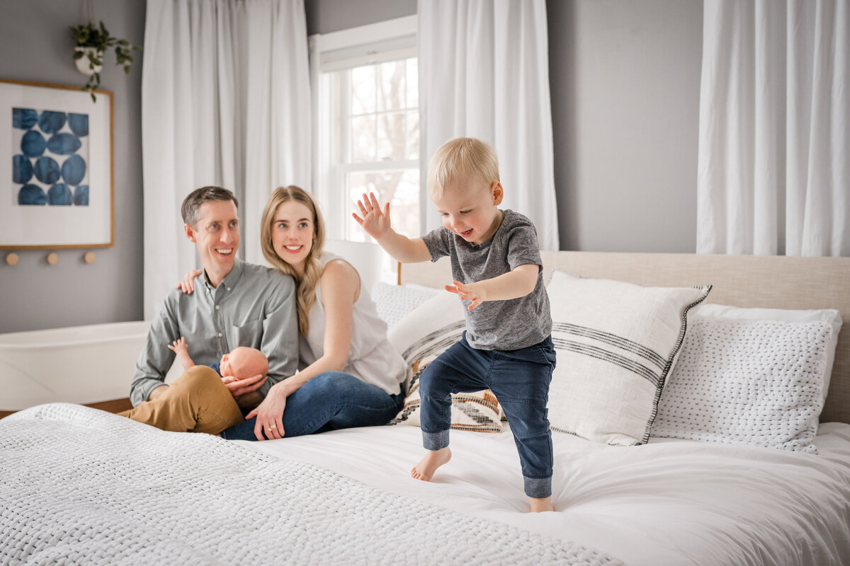A joyful toddler dances on a bed while his parents, sitting nearby with a newborn in their arms, smile and watch.