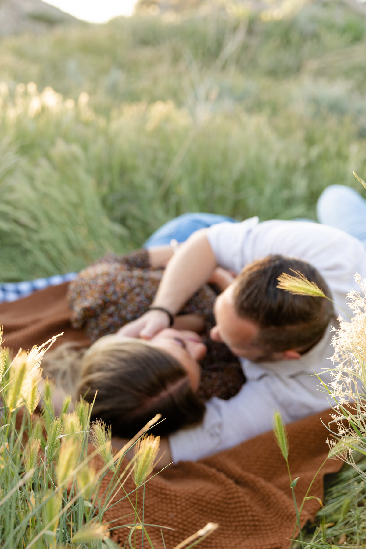 A couple lies together on a cozy brown blanket in a lush green meadow in Saratoga Springs, Utah. The man tenderly cradles the woman’s face as they share a romantic gaze, surrounded by tall wild grasses and soft golden sunlight
