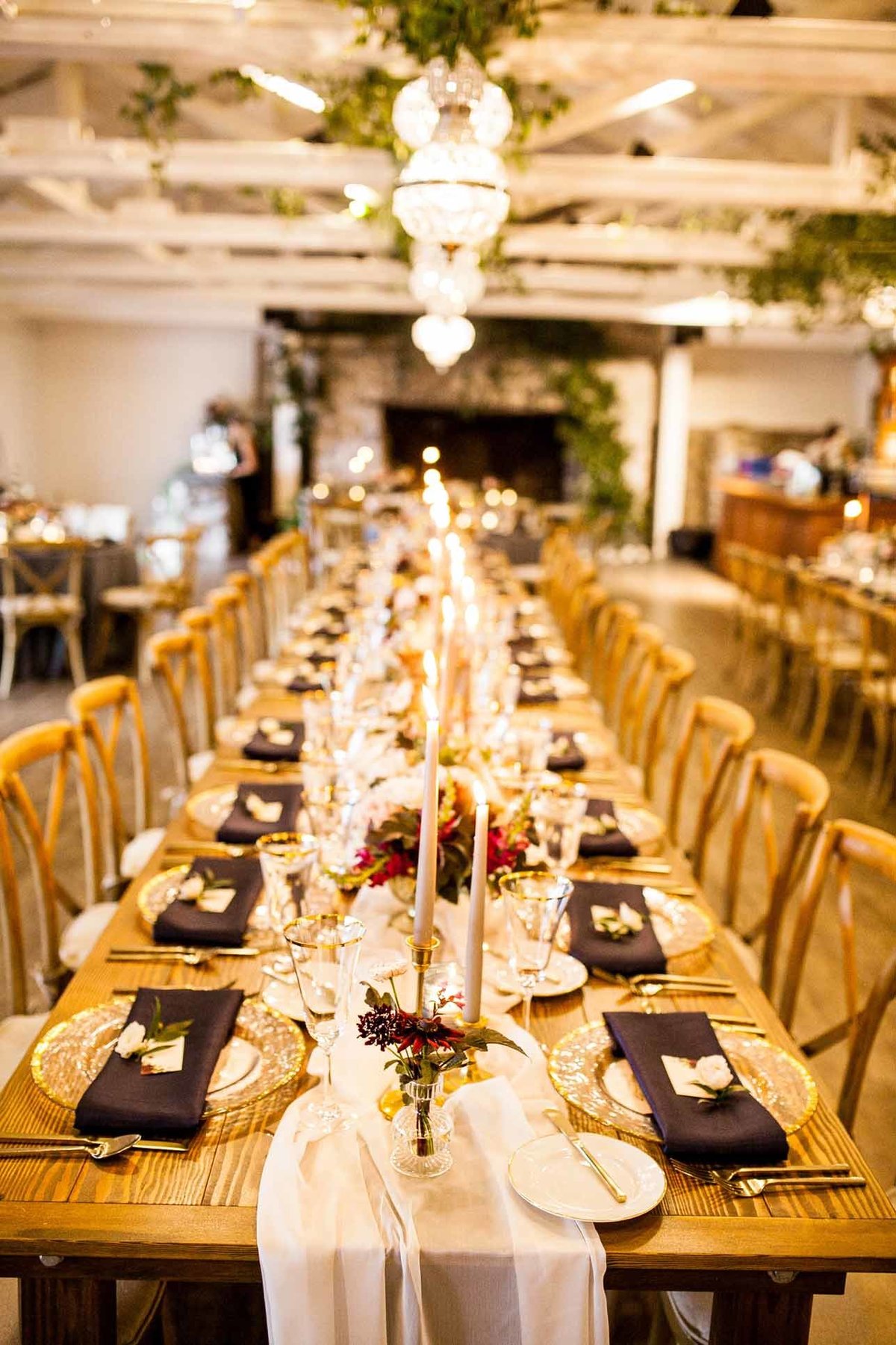 Long wooden tables with Vineyard chairs, gold rimmed glass chargers, ivory sheer table runner, and fall flowers