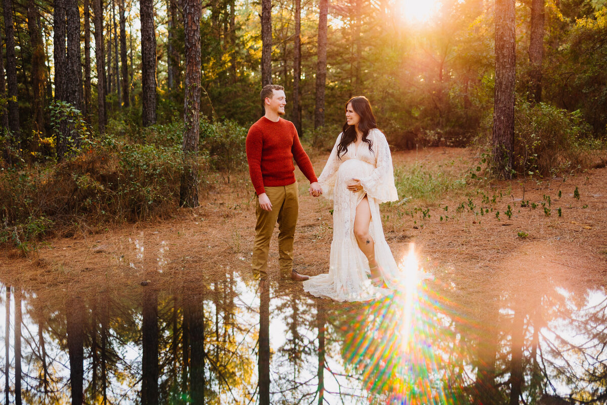 Professional maternity photography in Albuquerque capturing a new family by a serene lake in a lush forest. The family is dressed in vintage outfits, exuding happiness and warmth as they enjoy the picturesque setting