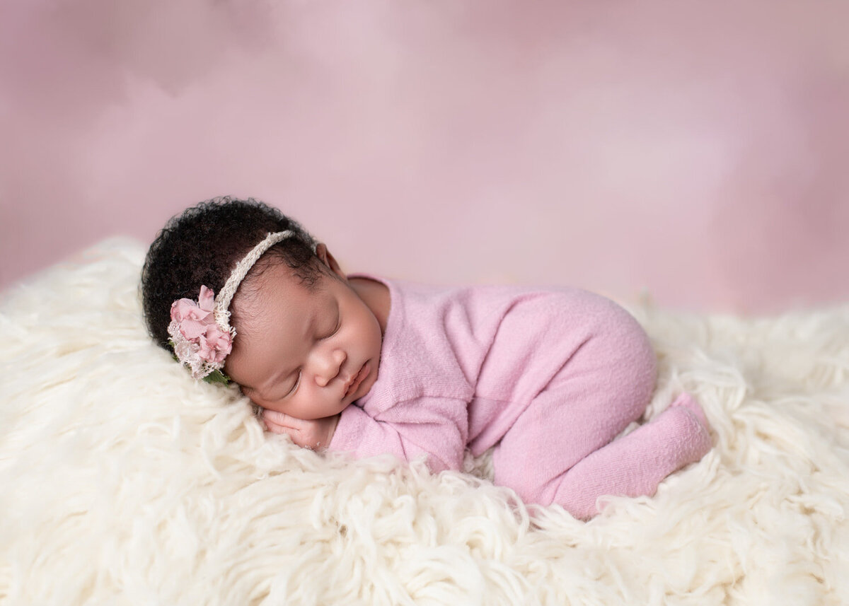 Newborn photography pricing — baby girl in pink sleeper with floral headband, posed on soft white flokati blanket