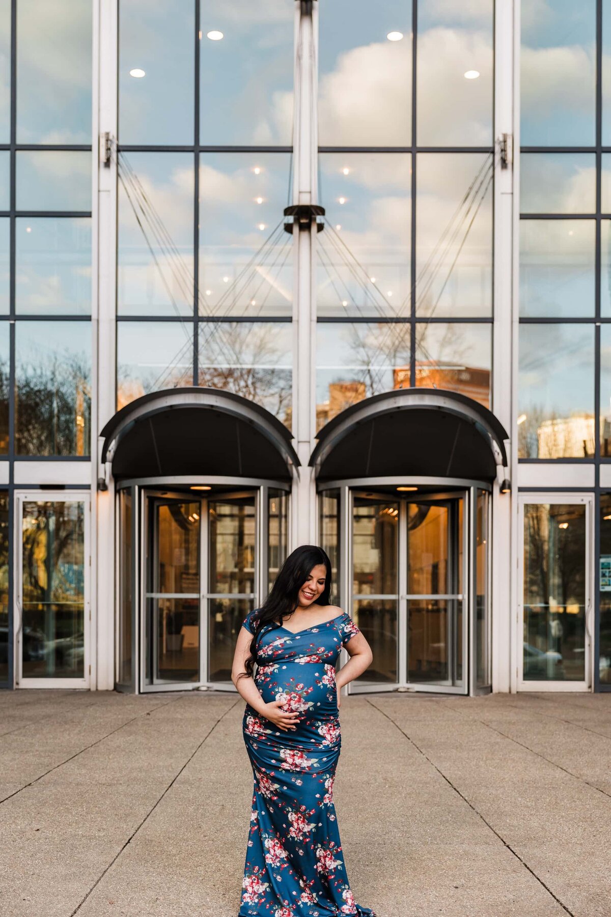 A smiling woman in a floral dress, captured by a Pittsburgh maternity photographer, stands in front of a building with glass doors.