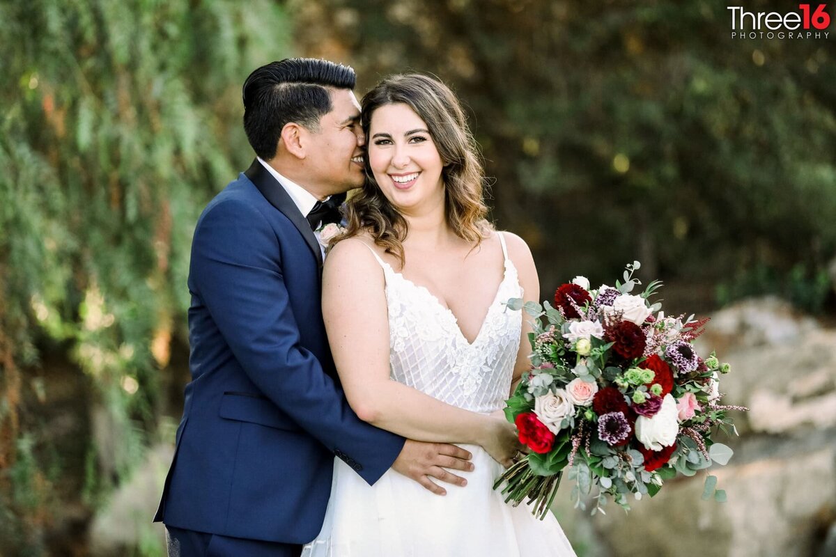 Groom hugs his Bride and whispers into her ear as the Bride gives a huge smile