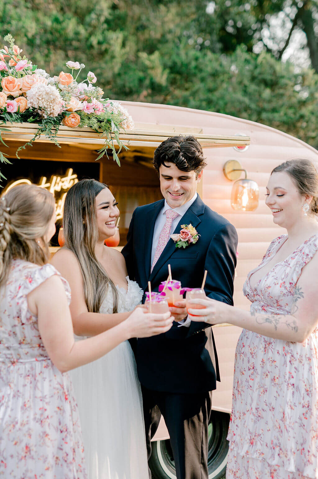 Bride and groom toast with friends in front of floral-decorated mobile bar at wedding reception.