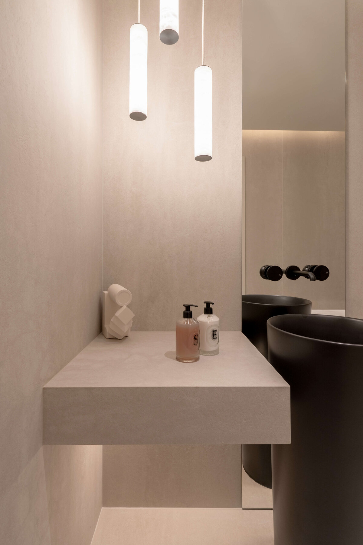 Modern bathroom featuring a light, airy aesthetic contrasted by a dramatic black pedestal sink, Three cylindrical pendant lights emit a warm glow.