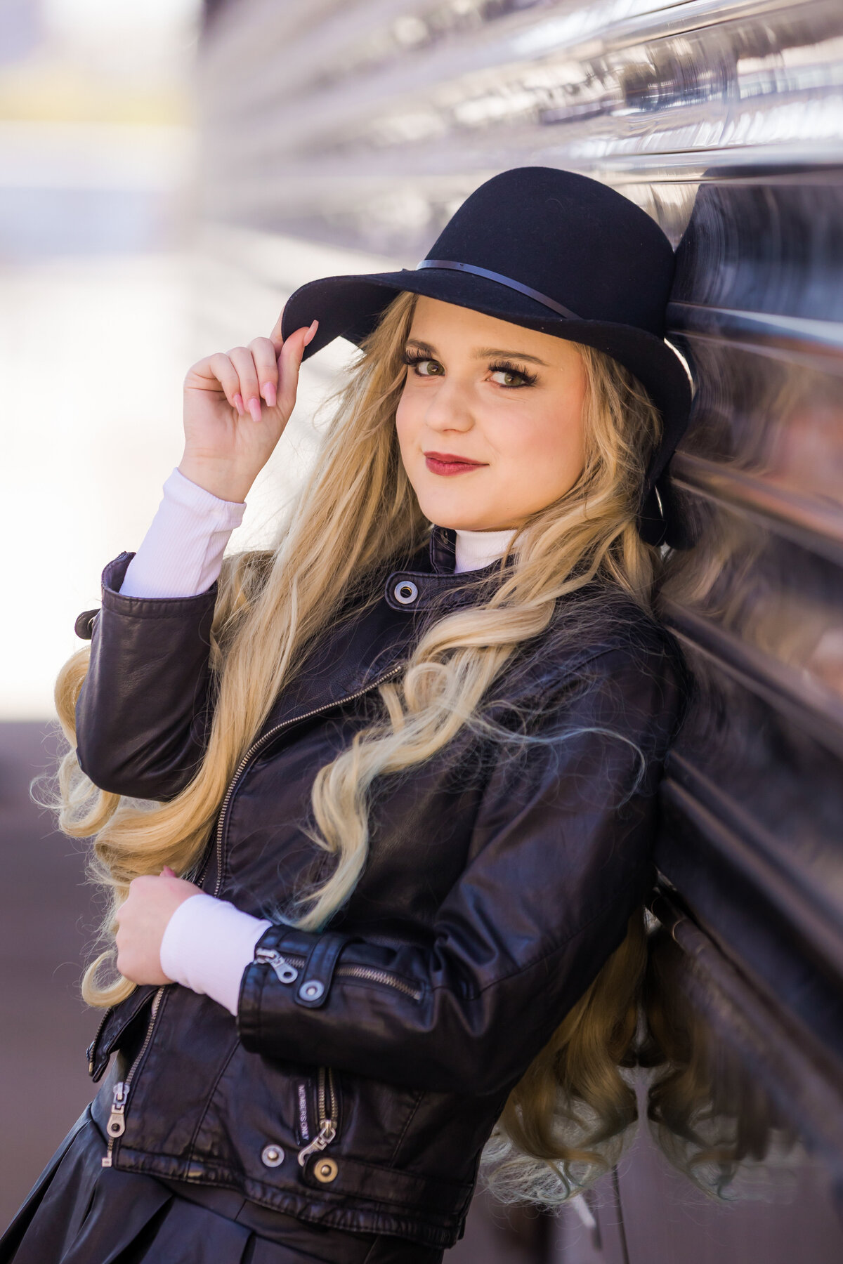 A teen girl holds her jacket with one hat while holding the edge of her hat with the other as she leans against a reflective wall.