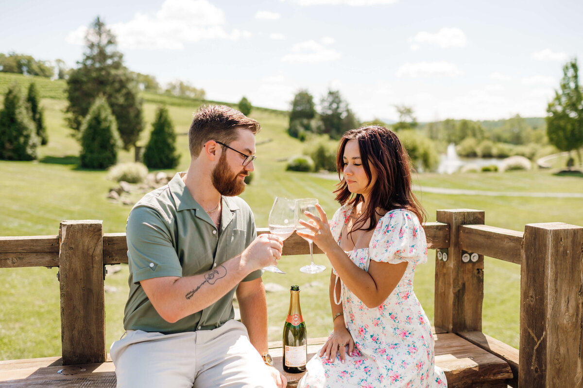 man and woman toasting champagne glasses together at a vineyard for summer engagement pictures