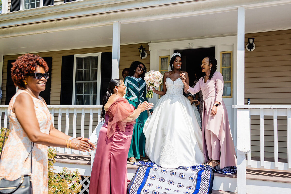 A joyous wedding scene on a sunny day with a bride in a white gown surrounded by smiling bridesmaids in pink dresses on a porch, while a lady in a floral dress walks by.