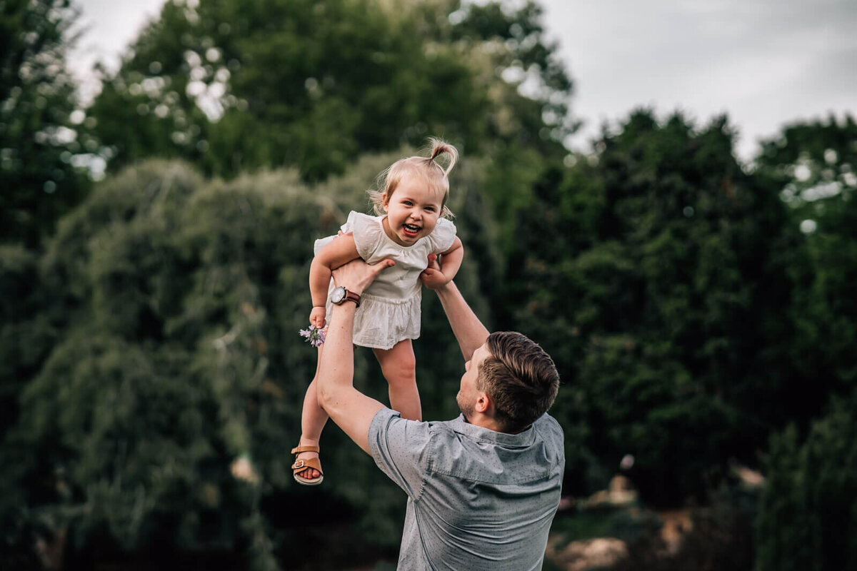 A young father gently throws his toddler into the air, while she facing the camera , laughing.