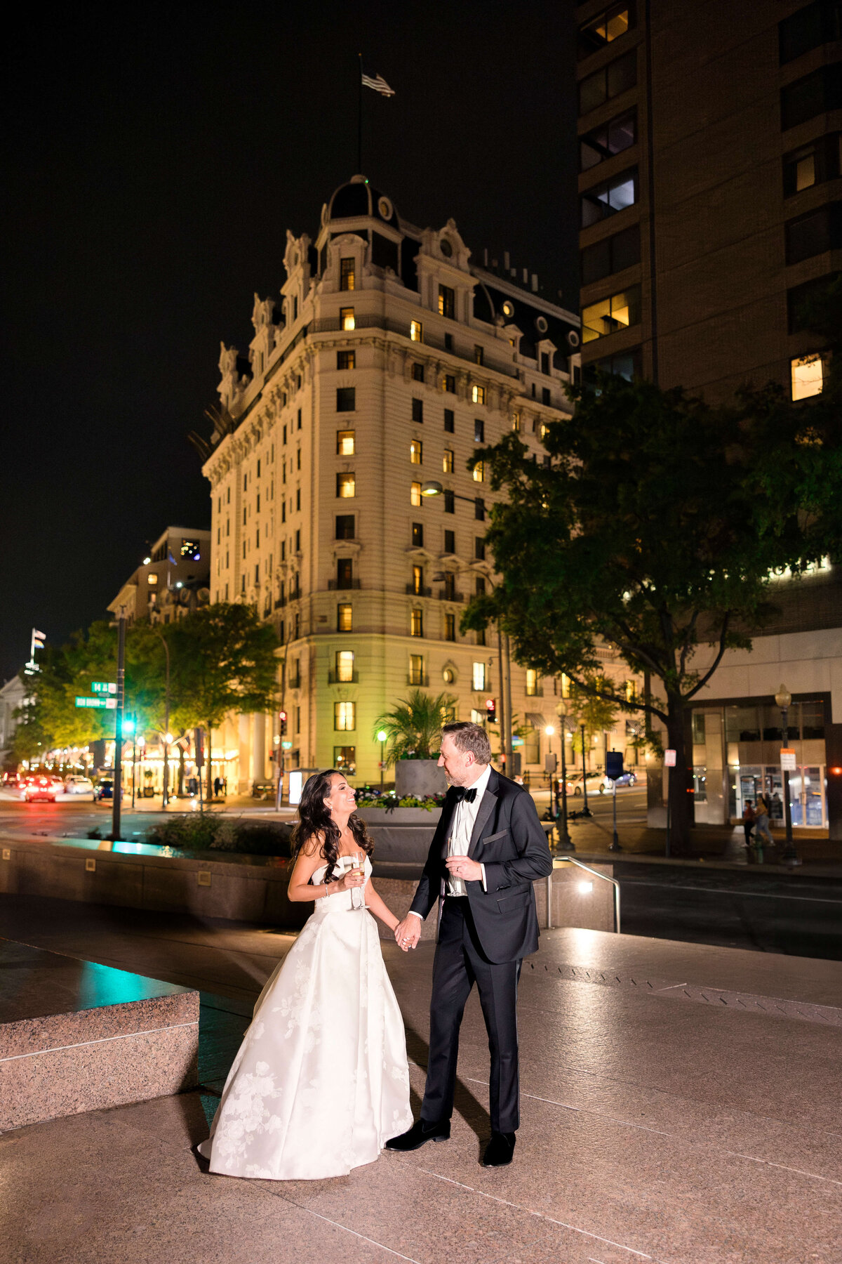 A couple in formal attire stands outside at night. The woman wears a white gown, and the man wears a black suit. They are holding hands and smiling. City lights illuminate the scene, highlighting a historic building in the background.