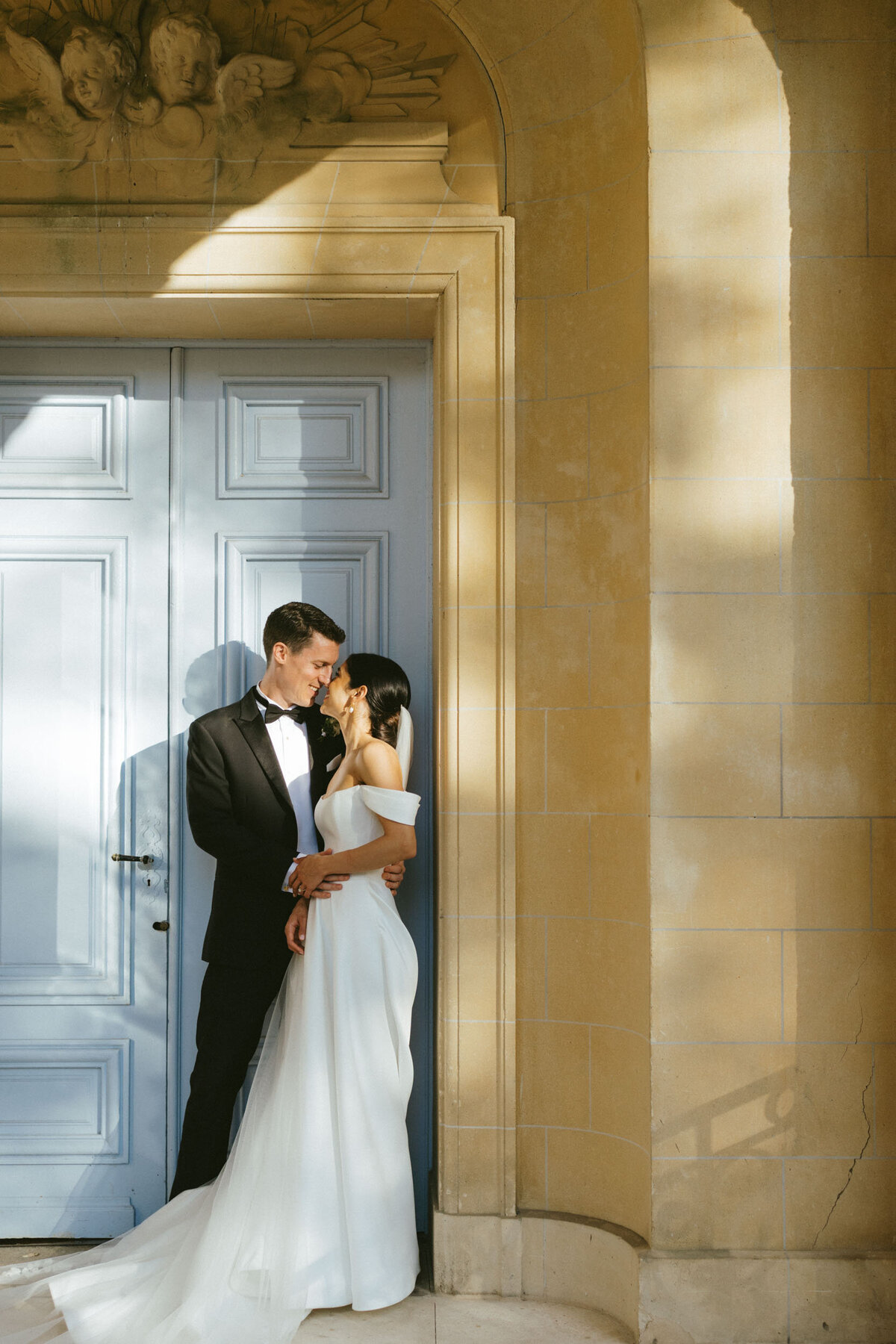 A bride and groom embrace intimately in a sunlit historic chateau in Paris, France.