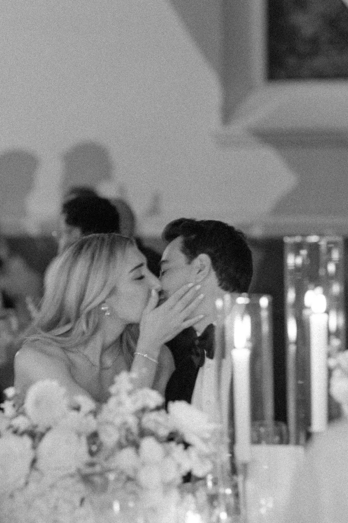 Couple de mariés s'embrassent tendrement lors de la réception de leur mariage, devant eux, une table remplie de fleurs et des chandelles. 