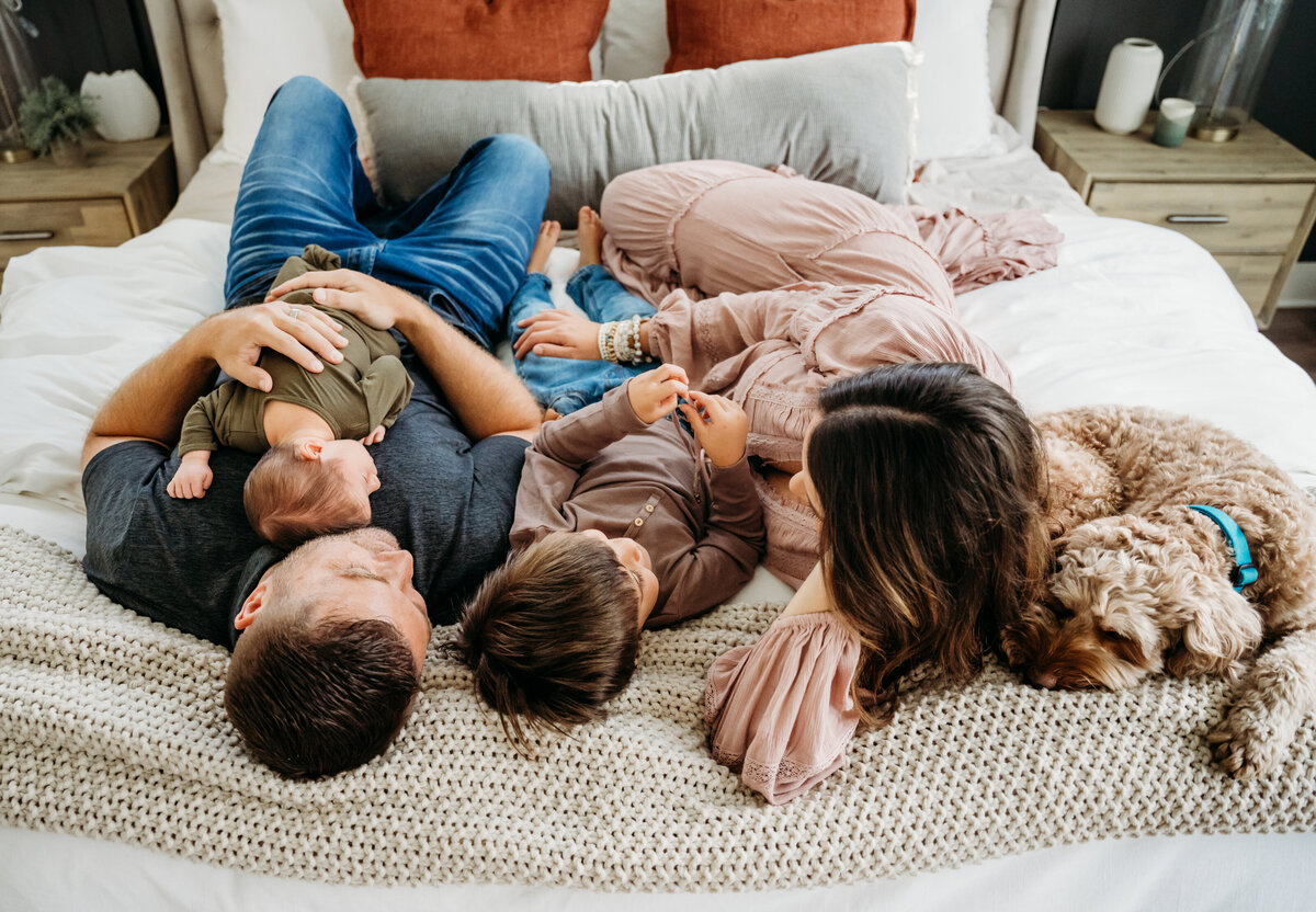 Newborn Photographer, a young family of four lays on the bed with their dog, newborn baby lays on dad's back