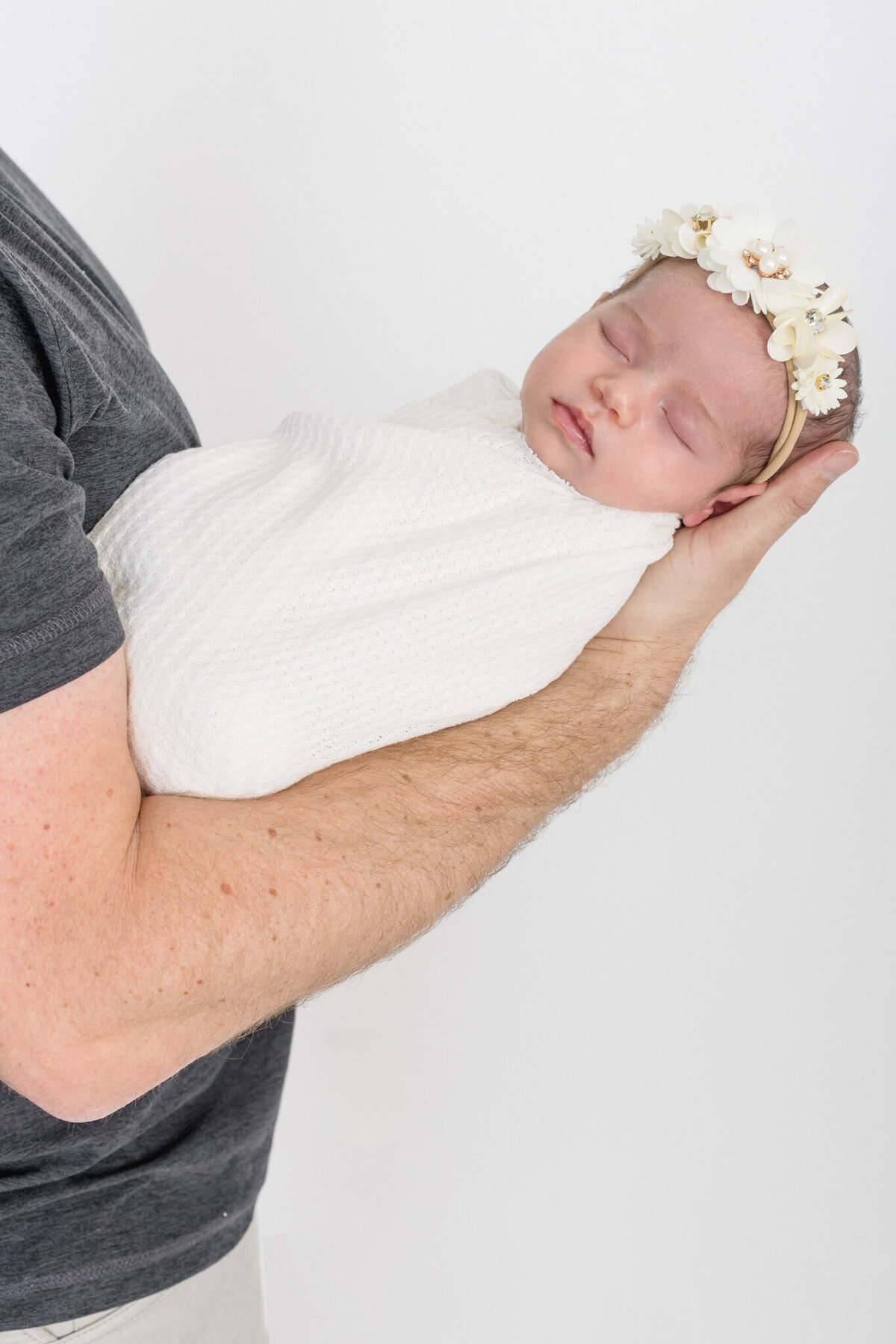 A baby wrapped in a white blanket sleeps peacefully, held by an adults arm. The baby wears a floral headband with small white flowers. The background is plain white.