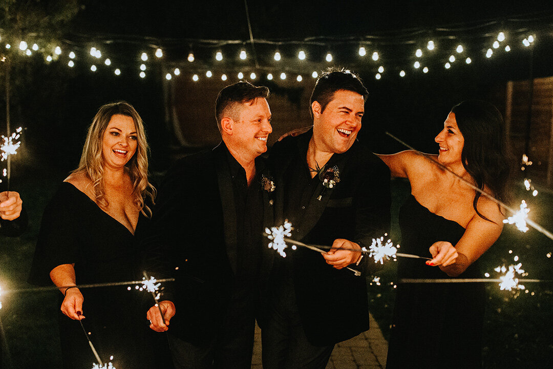 Two grooms wearing black tuxedos hold light-up sparklers standing next to two women wearing black wedding gowns at night.